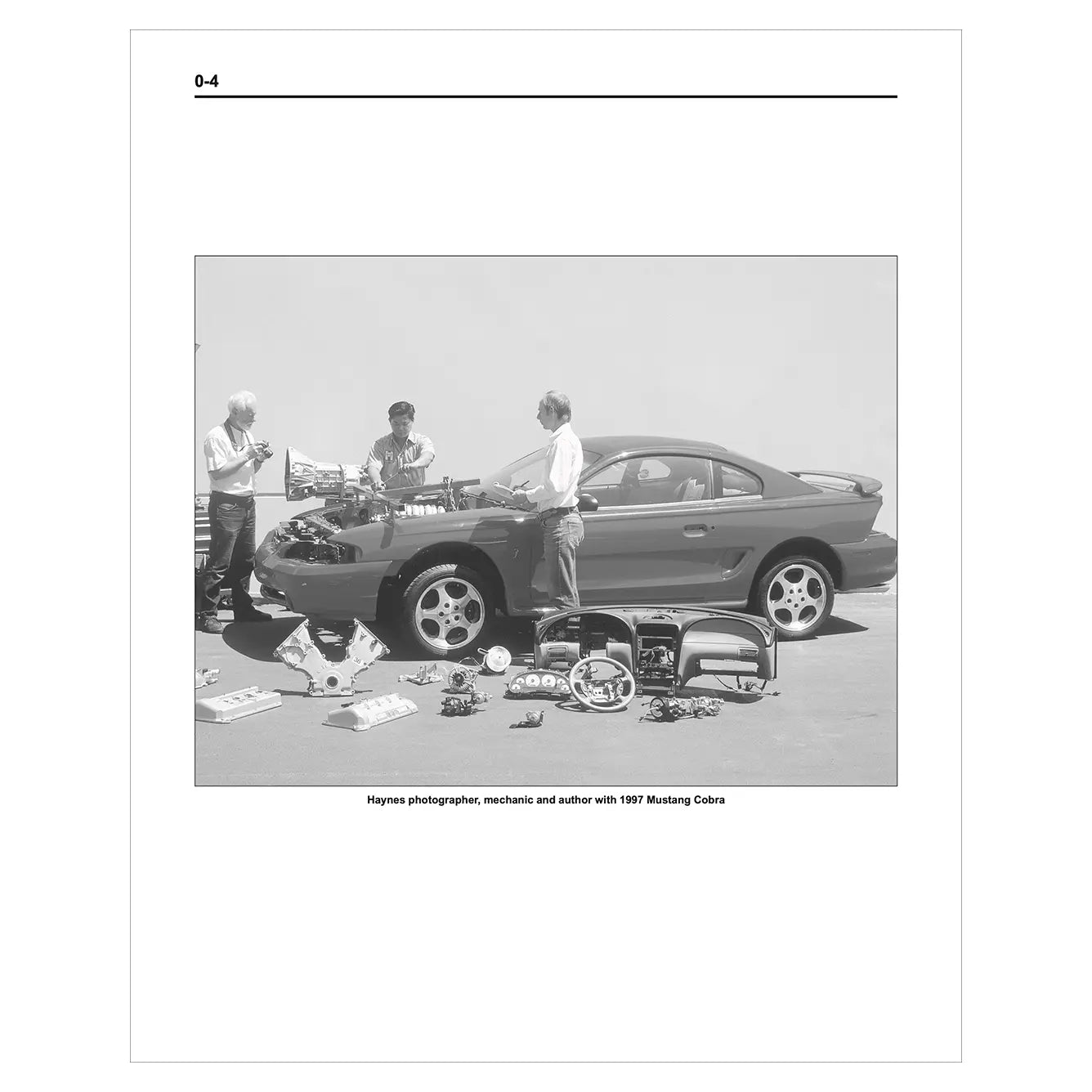 Black and white photo of a car with parts on a table and people around it.